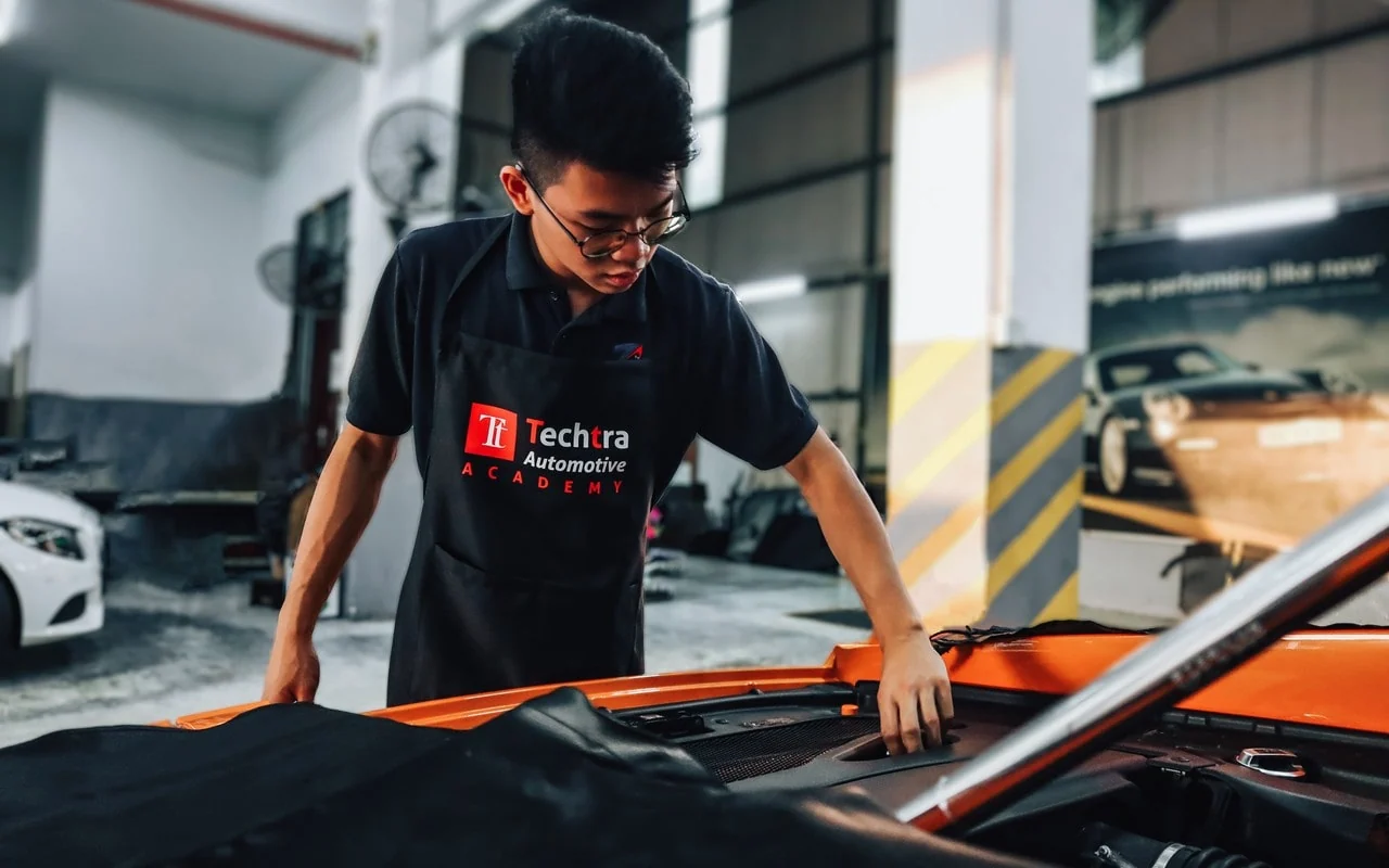 A person wearing a Techtra Automotive Academy apron examines an orange vehicle in a workshop